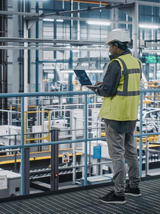 a man in a safety vest and hardhat using a laptop looking over manufacturing plant